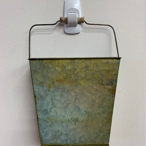 May include: A galvanized metal bucket with a wire handle and a wooden grip, hanging on a white wall. The bucket has a weathered, mottled appearance with shades of green and gold.