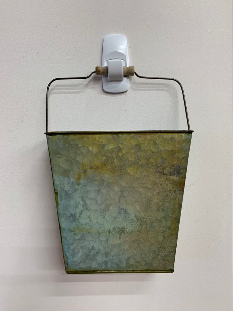May include: A galvanized metal bucket with a wire handle and a wooden grip, hanging on a white wall. The bucket has a weathered, mottled appearance with shades of green and gold.