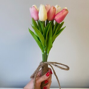 May include: A bouquet of artificial tulips in shades of pink, white, and cream, with green leaves. The stems are bound with twine, tied in a bow. The flowers are held in a hand with red nail polish.