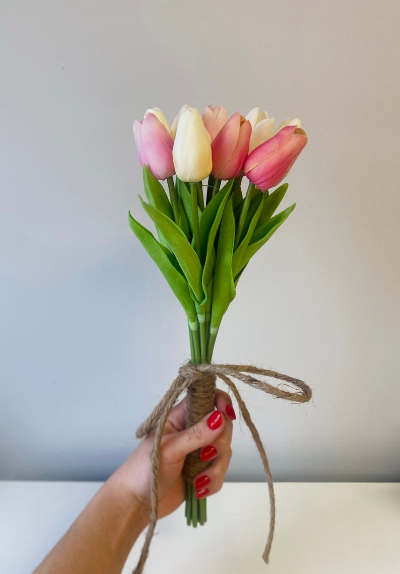 May include: A bouquet of artificial tulips in shades of pink, white, and cream, with green leaves. The stems are bound with twine, tied in a bow. The flowers are held in a hand with red nail polish.