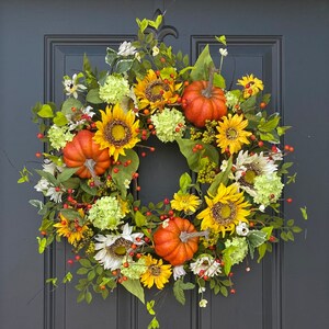 May include: A vibrant autumn wreath featuring sunflowers, orange pumpkins, white and yellow flowers, green hydrangeas, and red berries. The wreath is set against a dark gray door, creating a warm and inviting seasonal decoration.