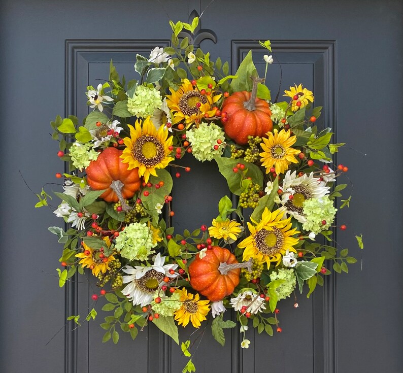 May include: A vibrant autumn wreath featuring sunflowers, orange pumpkins, white and yellow flowers, green hydrangeas, and red berries. The wreath is set against a dark gray door, creating a warm and inviting seasonal decoration.