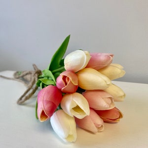 May include: A bouquet of artificial tulips in shades of pink, cream, and yellow, tied with twine. The flowers have green leaves and are arranged on a white surface against a gray background. The tulips are a popular choice for home decor.