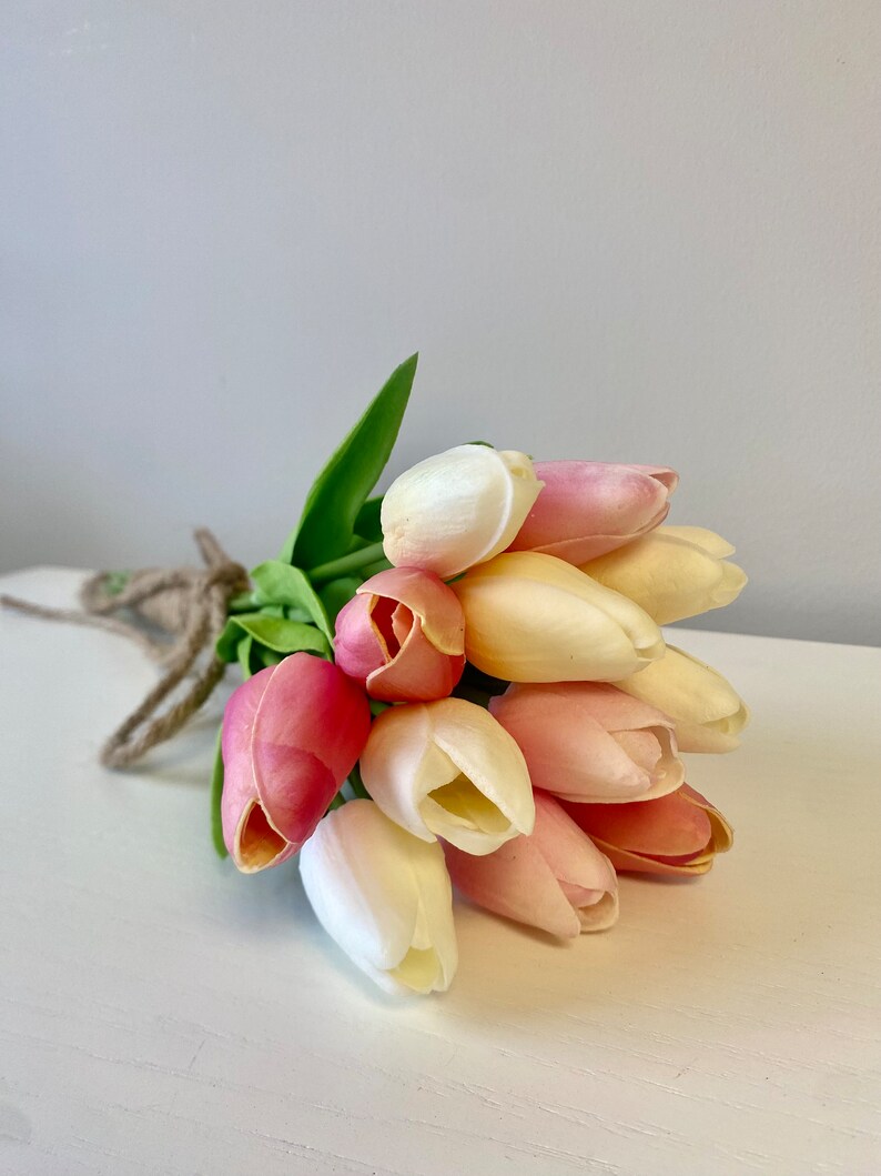 May include: A bouquet of artificial tulips in shades of pink, cream, and yellow, tied with twine. The flowers have green leaves and are arranged on a white surface against a gray background. The tulips are a popular choice for home decor.