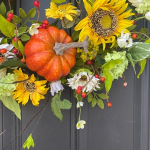 May include: A fall-themed wreath featuring a vibrant orange pumpkin, sunflowers, white daisies, and green foliage. Red berries and green clusters add pops of color. The wreath is designed for seasonal home decor.