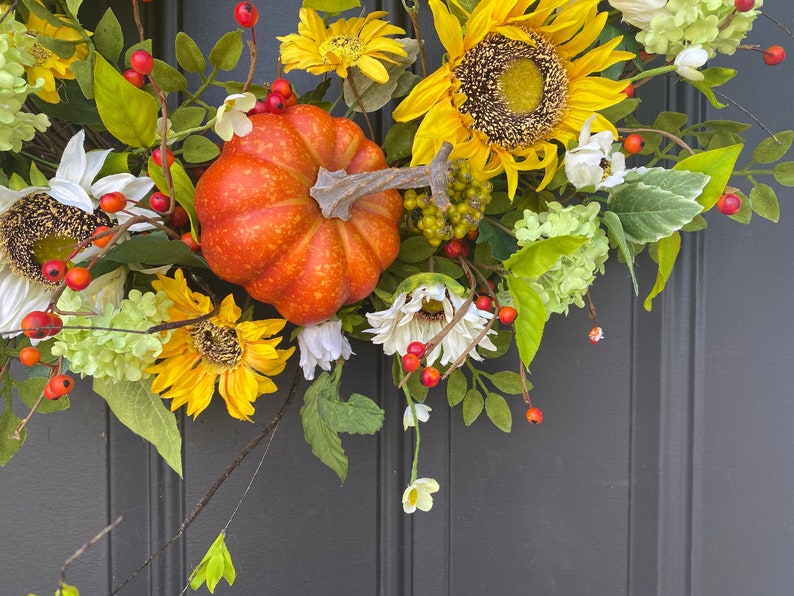 May include: A fall-themed wreath featuring a vibrant orange pumpkin, sunflowers, white daisies, and green foliage. Red berries and green clusters add pops of color. The wreath is designed for seasonal home decor.