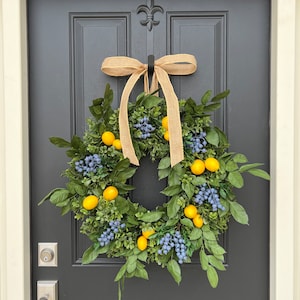 May include: A wreath made of green leaves and blue berries with yellow lemons hanging from the branches. The wreath is tied with a brown burlap ribbon and is hanging on a black door.