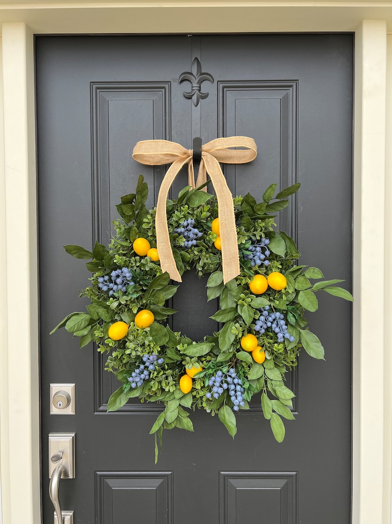 May include: A wreath made of green leaves and blue berries with yellow lemons hanging from the branches. The wreath is tied with a brown burlap ribbon and is hanging on a black door.