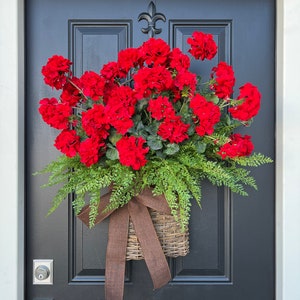 May include: A red geranium flower arrangement in a brown wicker basket with a brown bow, hanging on a black door.
