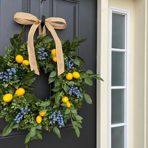 May include: A wreath made of green leaves and blue berries with yellow lemons hanging from it. The wreath is tied with a brown burlap ribbon and is hanging on a black door.