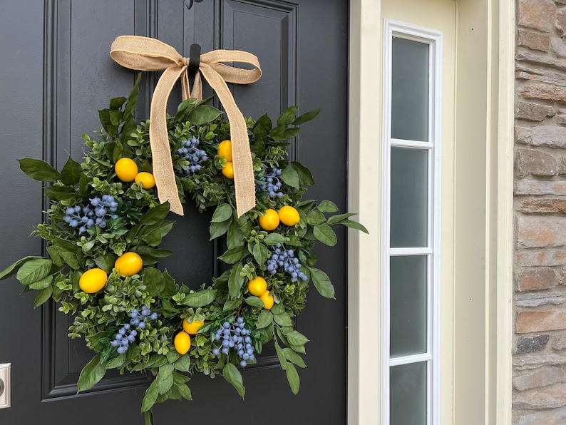 May include: A wreath made of green leaves and blue berries with yellow lemons hanging from it. The wreath is tied with a brown burlap ribbon and is hanging on a black door.