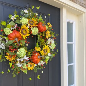 May include: A fall-themed wreath with sunflowers, orange pumpkins, white and green hydrangeas, and red berries. The wreath is displayed on a dark gray door, creating a festive autumn decoration.