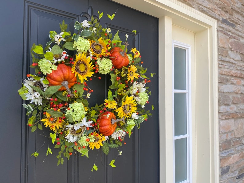 May include: A fall-themed wreath with sunflowers, orange pumpkins, white and green hydrangeas, and red berries. The wreath is displayed on a dark gray door, creating a festive autumn decoration.