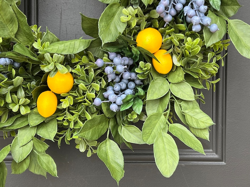May include: A green wreath with yellow lemons and blue berries. The wreath is hanging on a gray door.