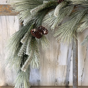 May include: A festive garland featuring long, green pine needles dusted with white frost. Brown pinecones are nestled among the branches. The garland is arranged on a rustic, wooden background, evoking a winter holiday theme.