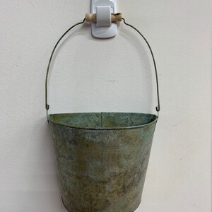 May include: A small, weathered metal bucket with a wire handle and a wooden grip. The bucket has a distressed, aged appearance with shades of green and brown. It is hanging on a white wall.
