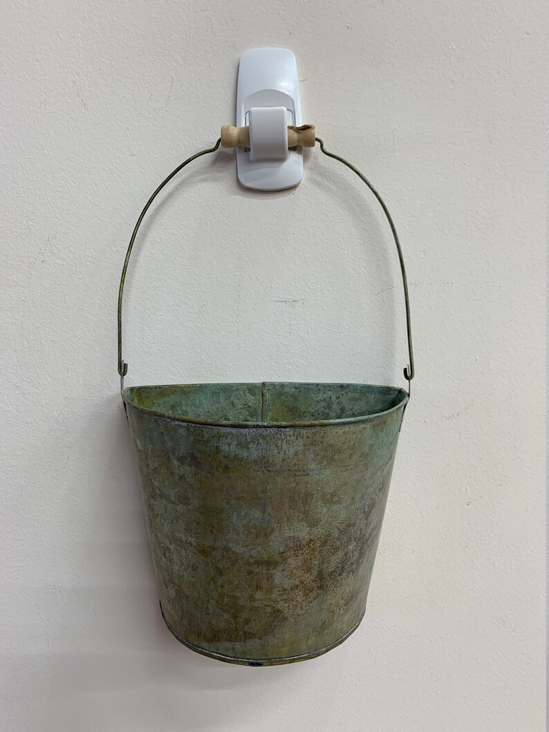 May include: A small, weathered metal bucket with a wire handle and a wooden grip. The bucket has a distressed, aged appearance with shades of green and brown. It is hanging on a white wall.