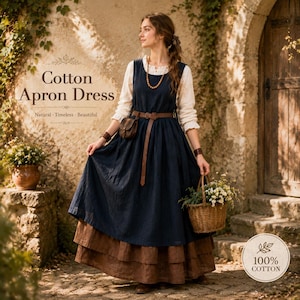 May include: A dark blue cotton apron dress worn over a long-sleeved cream-colored blouse. The dress features a brown leather belt at the waist, a full-length skirt with a brown tiered hem, and is paired with a small brown leather pouch.
