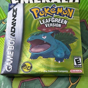 May include: A sealed Pokémon LeafGreen Version game cartridge for Game Boy Advance. The box features a green and gold color scheme with the Pokémon character, Venusaur, prominently displayed. The game is rated "Everyone" by the ESRB.