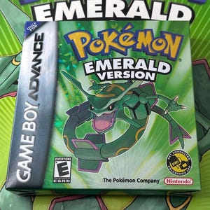 May include: A Game Boy Advance video game cartridge box for Pokémon Emerald Version. The box is green with the Pokémon logo and a picture of a green dragon-like creature. The box has the ESRB rating and the Nintendo logo.