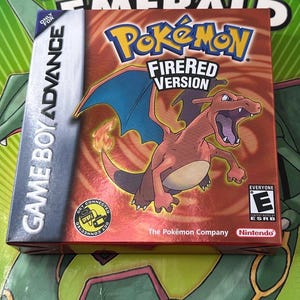 May include: A Game Boy Advance video game box for Pokémon FireRed Version. The box features a red and orange background with a Charizard illustration. The Pokémon logo is in yellow and blue. The ESRB rating is visible.