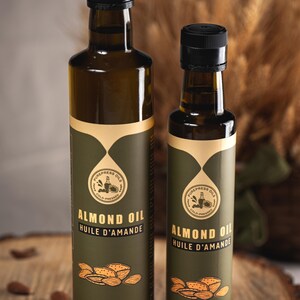 May include: Two glass bottles of almond oil, one 500 ml and one 250 ml, with green labels and gold accents. The labels feature a logo with the text "PUREPRESS OILS COLD-PRESSED" and the words "ALMOND OIL HUILE D'AMANDE".