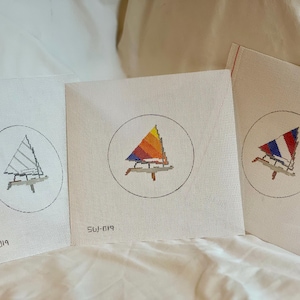 May include: Three white canvas needlepoint kits with different sailboat designs. The first kit features a black and white sailboat, the second kit features a red, orange, and yellow sailboat, and the third kit features a red, white, and blue sailboat. Each kit has a circle around the sailboat design and the text "SW-019" in the bottom left corner.
