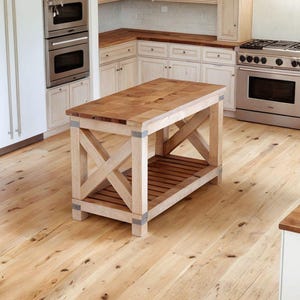 Pode incluir: Uma cozinha iluminada com armários de madeira clara, um forno e fogão de aço inoxidável e uma grande ilha de madeira. A ilha tem uma base em forma de X e um tampo de bloco de açougueiro. O chão é de madeira clara.