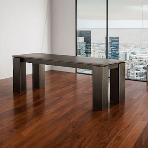 May include: A dark brown rectangular table with four square legs. The table is in a room with a large window overlooking a cityscape. The floor is made of dark wood.