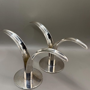 May include: Three silver-colored candle holders with a modern design. Each holder has a circular base and a curved, arching top. The polished metal reflects light, creating a sleek and elegant appearance. The candle holders are suitable for home decor.