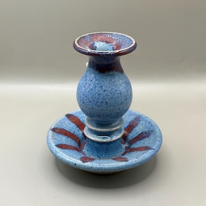 May include: A blue and red ceramic candle holder with a wide base and a tall, slender top. The holder has a speckled glaze and red accents.