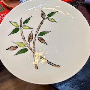 May include: A round, off-white ceramic plate with a hand-painted bamboo design. The bamboo stalks are tan with dark brown detailing, and the leaves are green and yellow. The plate has a slightly textured surface.