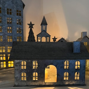 May include: A collection of metal houses and buildings with cut-out windows and doors. The structures are painted in shades of gray and have a rustic, weathered appearance. Some buildings have a star or tree on top, and the windows glow with warm light.