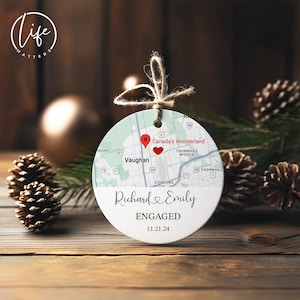 May include: A round, white ceramic ornament with a map design, a red heart, and a red location pin. The ornament has the names "Richard & Emily" and the word "ENGAGED" with the date "11.21.24". The ornament is hanging by a twine string.