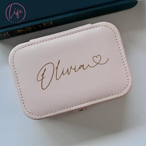 May include: A pale pink jewelry box with a gold inscription that reads "Olivia" with a small heart symbol.