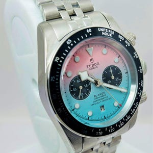May include: A stainless steel Blaken chronograph watch with a gradient pink and blue dial. The watch features a black bezel with white markings and a stainless steel bracelet. The dial includes sub-dials and the text "TUDOR GENEVE".