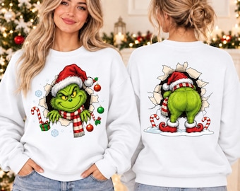 May include: A white long-sleeve sweatshirt featuring a graphic of a green character wearing a Santa hat and scarf. The front shows the character peeking through a torn paper effect with ornaments, while the back shows the character from behind.