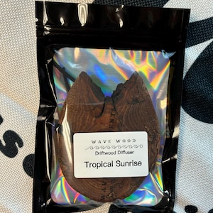 Puede incluir: Un difusor de madera flotante marrón en una bolsa de plástico transparente con una etiqueta blanca que dice "Wave Wood Driftwood Diffuser Tropical Sunrise".