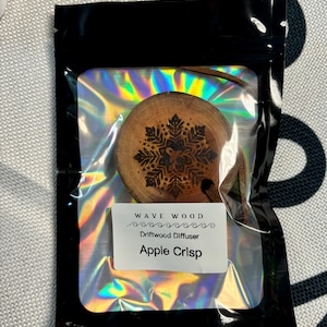 Op de afbeelding: Een zwarte pouch met een holografische, regenboogkleurige binnenkant. In de pouch zit een ronde houten diffuser met een sneeuwvlokkenpatroon. De diffuser heeft een label met de tekst "Wave Wood Driftwood Diffuser Apple Crisp".