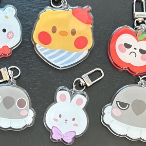 May include: A collection of six acrylic keychains featuring various cartoon characters, including a white round creature, a yellow character with a red beret, a red apple with a face, two grey birds with white ruffs, and a white rabbit with a purple bow.