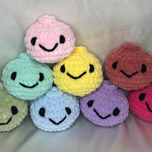 May include: A collection of eight round, crocheted plush toys in various colors including pink, mint green, olive green, light blue, yellow, purple, dark red, and hot pink. Each plush features two black embroidered eyes and a black stitched smile.
