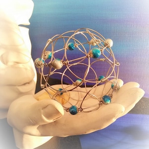 May include: A wire sphere sculpture with blue and white beads. The sphere is made of wire that is woven together to create a geometric pattern. The beads are evenly spaced around the sphere.