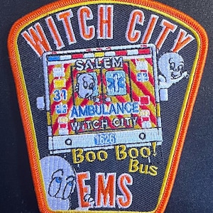 May include: Embroidered patch with the words "Witch City" in orange and white. The patch features an ambulance with ghost characters and the text "Boo Boo! Bus" and "EMS". The patch is black with an orange border.