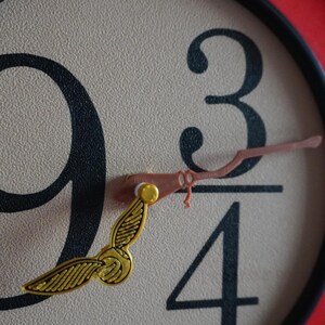 Puede incluir: Un reloj redondo con marco rojo y negro. La esfera es beige con números negros y un objeto alado dorado. La aguja de los minutos tiene forma de escoba. La hora es las nueve y cuarto.