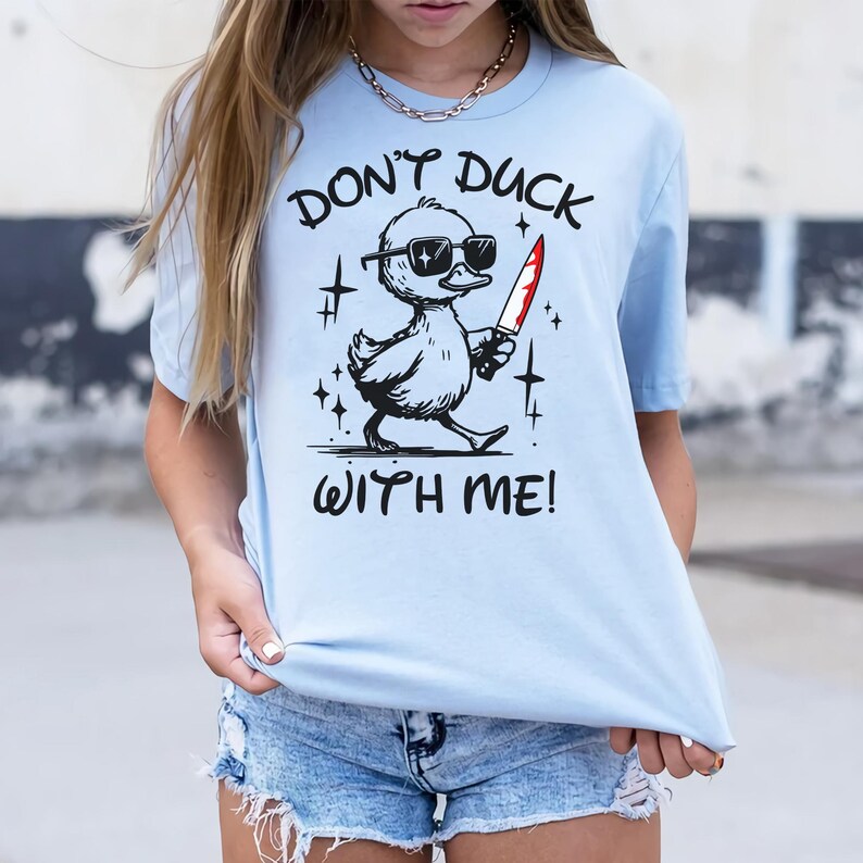 Pode incluir: Uma camiseta azul claro com um gr&aacute;fico de um pato de desenho animado usando &oacute;culos de sol e segurando uma faca ensanguentada. O texto "Don't duck with me!" est&aacute; impresso na camiseta.