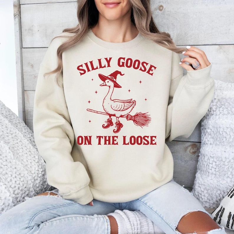 May include: Cream-colored sweatshirt with the text "SILLY GOOSE ON THE LOOSE" in red. The graphic features a goose wearing a witch hat and boots, riding a broom. The design is centered on the front of the long-sleeved top.