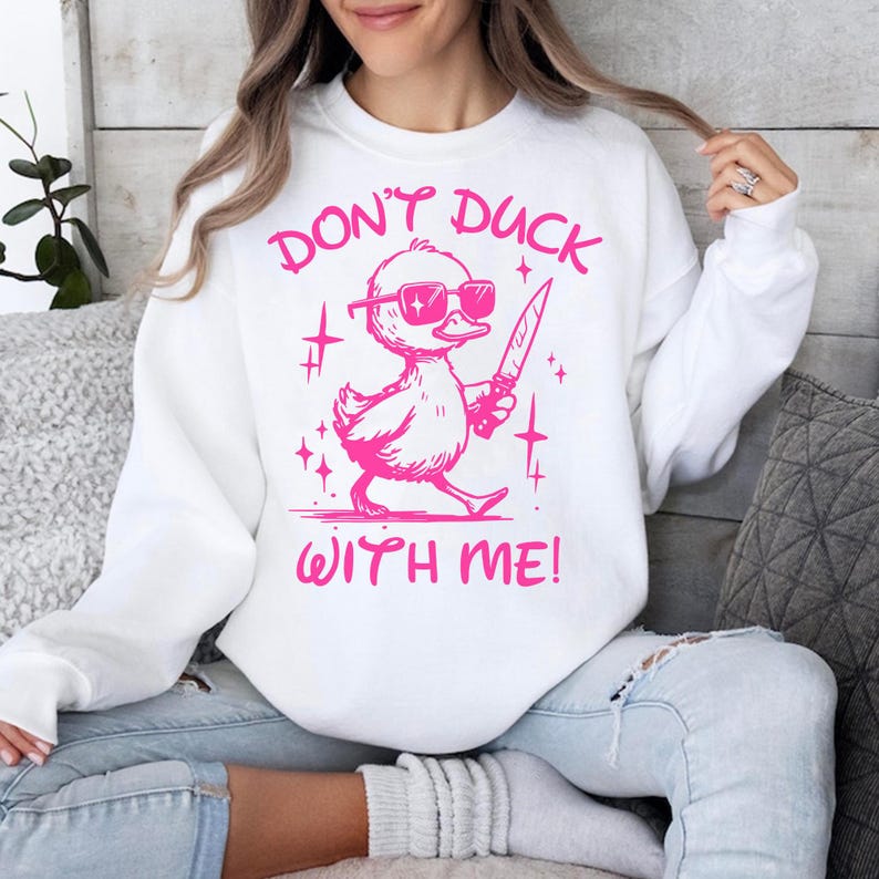 Pode incluir: Camisola branca com um gr&aacute;fico rosa de um pato usando &oacute;culos de sol e segurando uma faca. O texto "Don't Duck With Me!" est&aacute; impresso abaixo do pato.