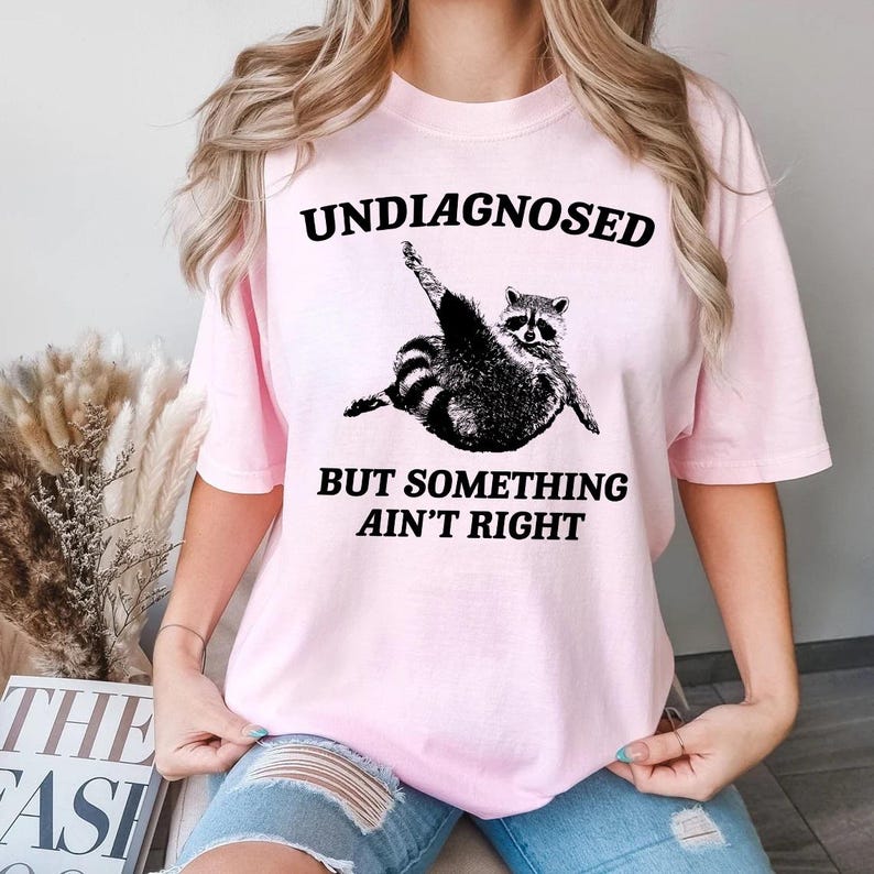 May include: Pink t-shirt with a black graphic of a raccoon doing yoga with the text "Undiagnosed but something ain't right"