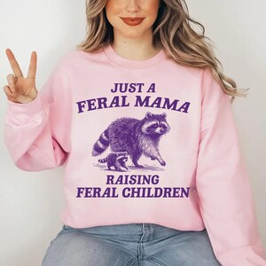 May include: Pink sweatshirt with a purple raccoon and baby raccoon graphic. The text reads "Just a Feral Mama Raising Feral Children."
