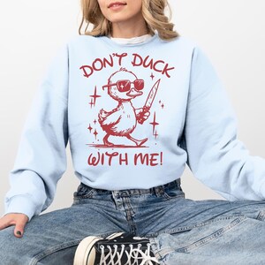 Pode incluir: Uma camisa de frio azul claro com o texto "Don't Duck With Me!" em vermelho. Um pato de desenho animado usando &oacute;culos de sol e segurando uma faca est&aacute; no centro do texto.